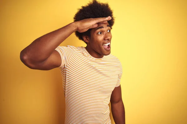 American man with afro hair wearing striped t-shirt standing over isolated yellow background very happy and smiling looking far away with hand over head. Searching concept.
