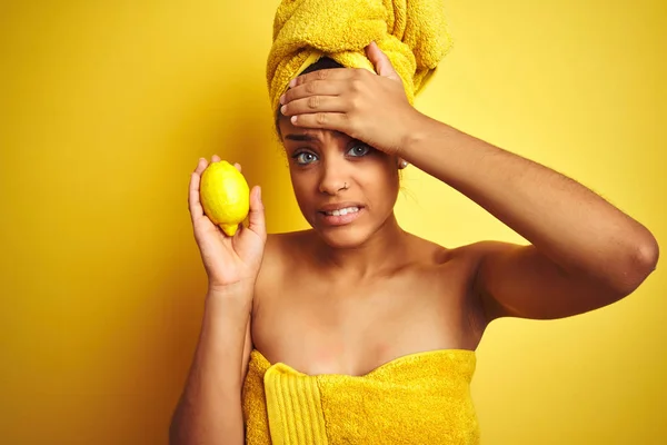 Afro american woman wearing towel after shower holding lemon over isolated yellow background stressed with hand on head, shocked with shame and surprise face, angry and frustrated. Fear and upset for mistake.