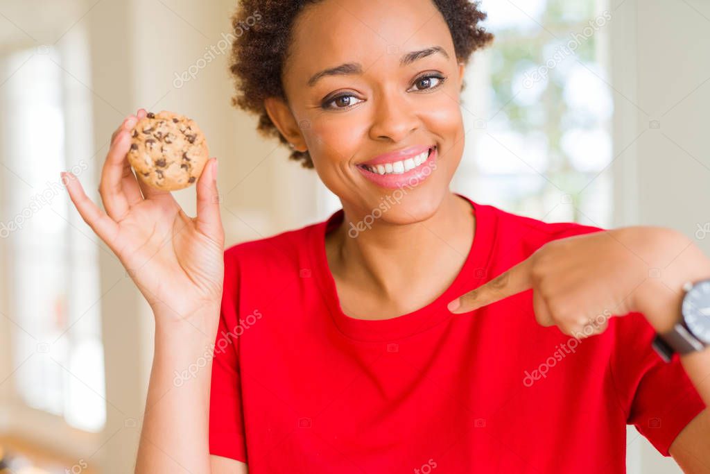 Young african american woman eating chocolate chips cookies with surprise face pointing finger to himself