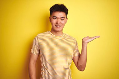 İzole sarı arka plan üzerinde duran t-shirt giyen genç Asyalı Çinli adam neşeli sunan gülümseyerek ve kameraya bakarak avuç içi ile işaret.