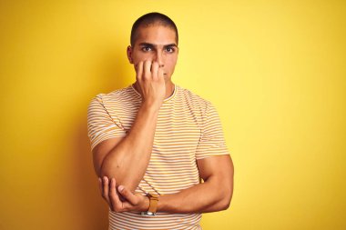 Genç yakışıklı adam sarı izole arka plan üzerinde çizgili t-shirt giyen stresli ve ağız tırnak ısırma elleri ile sinirli görünümlü. Anksiyete sorunu.