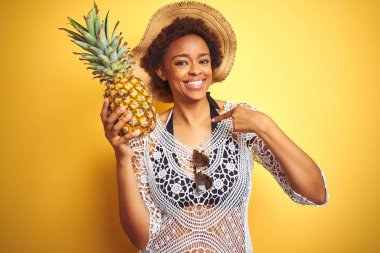 Kendine parmak işaret sürpriz yüz ile sarı izole arka plan üzerinde pineaple tutan afro saçlı genç afro-amerikalı kadın