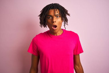 Izole pembe arka plan üzerinde duran t-shirt giyen dreadlocks giyen Afro Amerikan adam korkmuş ve sürpriz ifade, korku ve heyecanlı yüz ile şok.