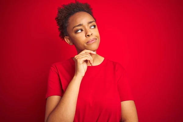Young beautiful african american woman with afro hair over isolated red background with hand on chin thinking about question, pensive expression. Smiling with thoughtful face. Doubt concept.
