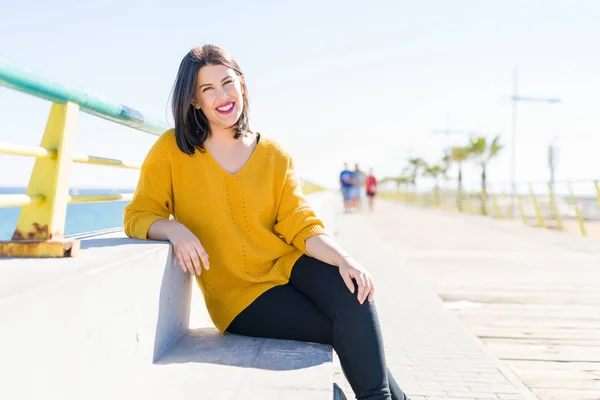 Mooie jonge vrouw zittend op een promenade door de zee glimlachend — Stockfoto