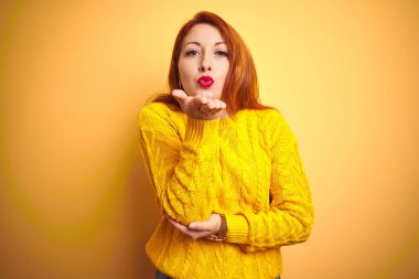 Güzel kızıl saçlı kadın izole sarı arka plan üzerinde duran kış kazak giyen kamera hava güzel ve seksi olmak el iyle bir öpücük üfleme bakarak. Aşk ifadesi.