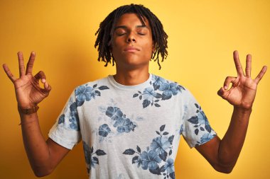 Izole sarı arka plan üzerinde yaz çiçek t-shirt giyen dreadlocks ile Afro adam rahatlayın ve parmakları ile meditasyon jest yapıyor kapalı gözleri ile gülümseyerek. Yoga konsepti.