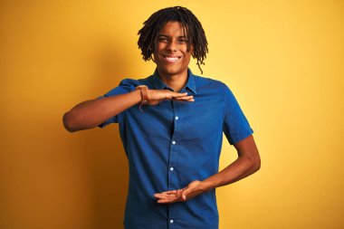 Dreadlocks rahat denim gömlek giyen afro adam elleri büyük ve büyük boy işareti gösteren izole sarı arka plan gesturing üzerinde duran, ölçü sembolü. Kameraya bakarak gülümsüyor. Ölçme kavramı.