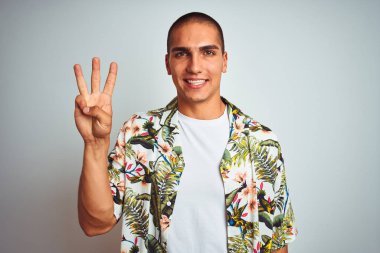 Genç yakışıklı adam tatillerde beyaz arka plan üzerinde Hawaii gömlek giyen gösteren ve emin ve mutlu gülümserken parmakları ile üç numara işaret.