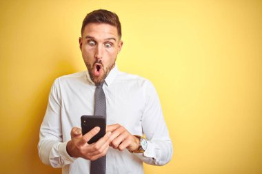 Genç yakışıklı iş adamı sarı izole arka plan üzerinde akıllı telefon kullanarak sürpriz bir yüz ile şok korkmuş, korku ifade korku ve heyecanlı