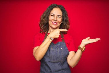 Kırmızı izole arka plan üzerinde önlük üniforma giyen orta yaş yaşlı kadın şaşırttı ve el ile sunarken kameraya gülümseyerek ve parmak ile işaret.