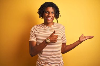 İzole sarı arka plan üzerinde duran çizgili t-shirt giyen dreadlocks ile Afro adam palmiye el gösteren ve başparmak ile ok jest yapıyor, mutlu ve neşeli gülümseyerek