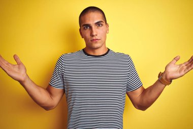 Genç yakışıklı adam sarı izole arka plan üzerinde çizgili t-shirt giyen habersiz ve kolları ve elleri kaldırdı ile karışık ifade. Şüphe kavramı.