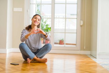 Evde oturan genç güzel kadın kalp sembolü ve elleri ile şekil gösteren aşk gülümsüyor. Romantik konsept.