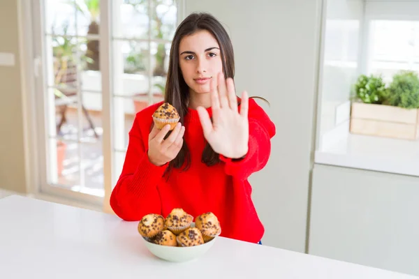 Beautiful young woman eating chocolate chips muffins with open hand doing stop sign with serious and confident expression, defense gesture
