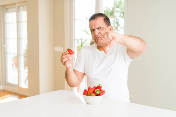 Middle age man eating strawberries at home with angry face, negative sign showing dislike with thumbs down, rejection concept