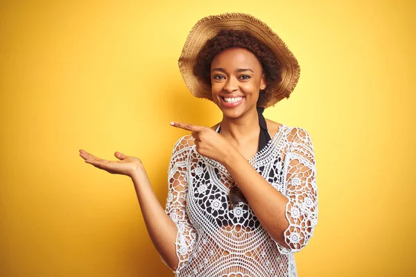 Young african american woman with afro hair wearing summer hat over white isolated background amazed and smiling to the camera while presenting with hand and pointing with finger.