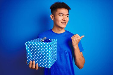 Genç asya çinli adam izole mavi arka plan üzerinde duran doğum günü hediyesi tutan işaret ve mutlu yüzü gülümseyerek yan başparmak ile gösteren