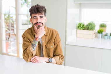 Emin çapraz kollar ve çeneme kaldırdı el ile gülümseme ile kameraya bakarak beyaz masada oturan rahat ceket genç adam. Pozitif düşünce.