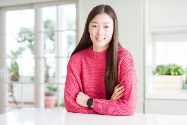 Beyaz masa üzerinde pembe kazak giyen güzel Asyalı kadın mutlu yüz kameraya bakarak çapraz kolları ile gülümseyerek. Pozitif kişi.