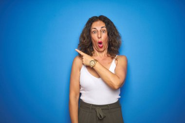 Kıvırcık saçlı orta yaş yaşlı kadın mavi izole arka plan üzerinde duran yan parmak ile işaret şaşırttı, açık ağız hayrete ifade.