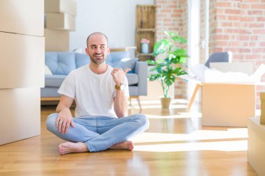 Genç kel adam karton kutular etrafında yerde oturan mutlu yüz görünümlü ve başparmak ile yan işaret gülümseyerek yeni bir eve hareket.