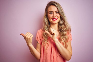 Pembe izole arka plan üzerinde duran t-shirt giyen genç güzel kadın el ve başparmak yukarı arka işaret, kendinden emin gülümseyerek