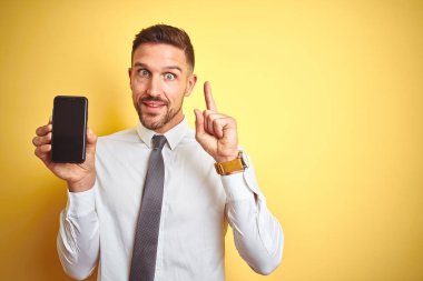 Genç yakışıklı iş adamı sarı izole arka plan üzerinde akıllı telefon ekranı gösteren mutlu yüz, bir numara ile bir fikir veya soru işaret parmağı ile şaşırttı