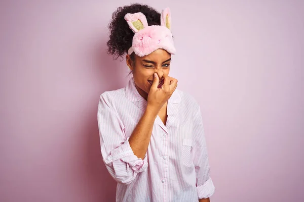 孤立したピンクの背景の上にパジャマとマスクを身に着けている若いアフリカ系アメリカ人女性は 鼻に指で息を保持し 臭いと嫌な 耐えぎにくい匂いを嗅ぐ 悪臭の概念 — ストック写真
