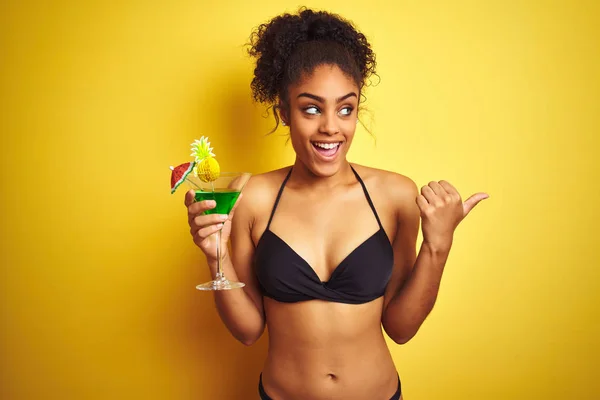 Afro woman on vacation wearing bikini drinking a cocktail over isolated yellow background pointing and showing with thumb up to the side with happy face smiling