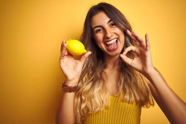 Young beautiful woman eating a lemon over yellow isolated background doing ok sign with fingers, excellent symbol