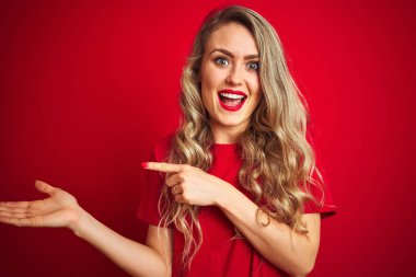 Kırmızı izole arka plan üzerinde duran temel t-shirt giyen genç güzel kadın şaşırttı ve el ile sunarken kameraya gülümseyerek ve parmak ile işaret.