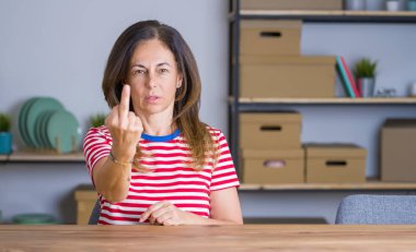 Orta yaşlı bir kadın evde masada oturuyor orta parmağını gösteriyor, kaba ve kaba bir ifade takınıyor.