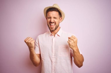 Yakışıklı adam pembe izole arka plan üzerinde şapka ve yazlık gömlek giyen çok mutlu ve heyecanlı kolları kaldırdı ile kazanan jest yapıyor, gülümseyerek ve başarı için çığlık. Kutlama konsepti.