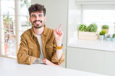 Yüzünde büyük bir gülümseme ile beyaz masanın üzerinde oturan rahat ceket giyen genç adam kameraya bakarak yan el ve parmak ile işaret.