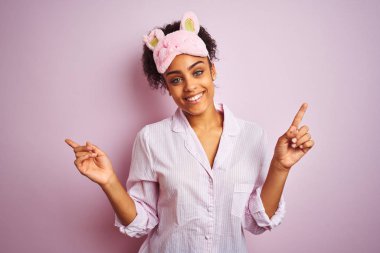 Genç afro-amerikan kadın izole pembe arka plan üzerinde pijama ve maske giyen farklı yönlere parmakları ile işaret emin gülümseyerek. Reklam için kopya alanı
