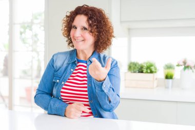 Evde kot ceket giyen kıvırcık saçlı orta yaş yaşlı kadın Beckoning mutlu ve gülümseyen davet eden el ile buraya jest geliyor