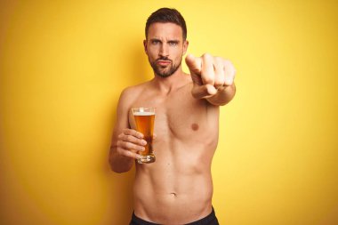 Genç yakışıklı gömleksiz adam kameraya parmak ile işaret izole sarı arka plan üzerinde bir bira bira içme ve size, el işareti, önden olumlu ve kendinden emin jest