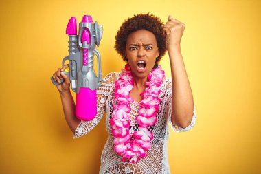 Afro saç giyen çiçek Hawaiian Lei ve su tabancası ile genç Afrikalı Amerikalı kadın rahatsız ve öfke, deli ve yükseltilmiş el, öfke kavramı ile bağırarak bağırıyor sinirli