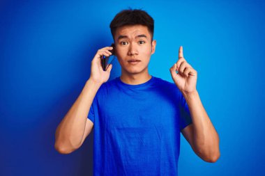 İzole mavi arka plan üzerinde duran akıllı telefon konuşan genç Asyalı Çinli adam mutlu yüz, bir numara ile bir fikir ya da soru işaret parmağı ile şaşırttı