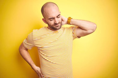 Sakallı genç kel adam sarı izole arka plan üzerinde rahat çizgili t-shirt giyen boyun ağrısı yaralanma acı, el ile boyun dokunmadan, kas ağrısı
