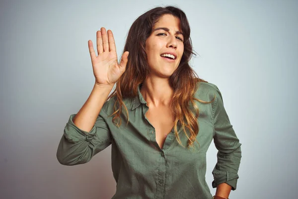 Young beautiful woman wearing green shirt standing over grey isolated background Waiving saying hello happy and smiling, friendly welcome gesture