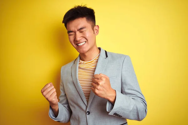 Young asian chinese businessman wearing jacket standing over isolated yellow background very happy and excited doing winner gesture with arms raised, smiling and screaming for success. Celebration concept.