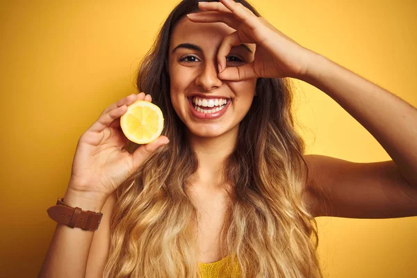 黄色の孤立した背景の上に半分のレモンを保持している若い美しい女性は 指を通して目に手でOkサインをやって笑顔で幸せな顔をしています — ストック写真