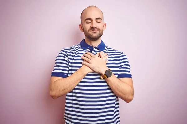 Young bald man with beard wearing casual striped blue t-shirt over pink isolated background smiling with hands on chest with closed eyes and grateful gesture on face. Health concept.