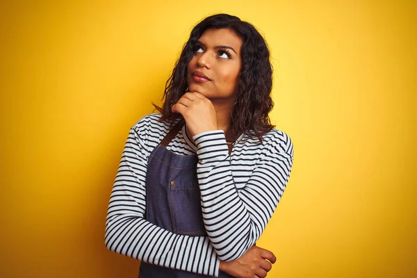 Genç transseksüel transseksüel şef kadın soru hakkında düşünme çene üzerinde el ile izole sarı arka plan üzerinde önlük giyen, dalgın ifade. Düşünceli bir yüzle gülümsüyor. Şüphe kavramı.