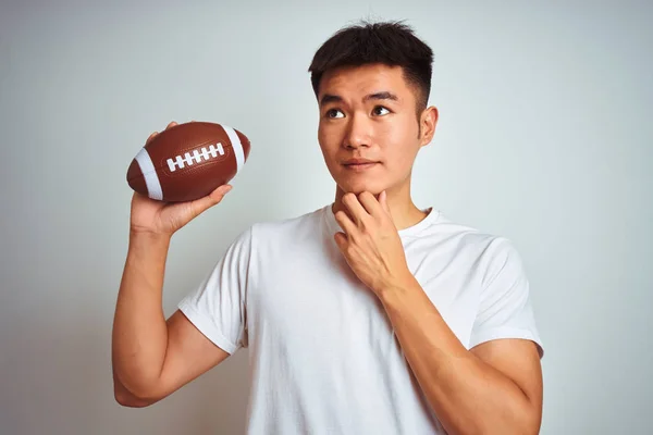 年轻的亚洲中国运动员拿着橄榄球站在孤立的白色背景严肃面对思考问题 很困惑的想法 — 图库照片