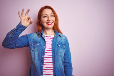 Denim gömlek giyen güzel kızıl saçlı kadın ve izole pembe arka plan üzerinde çizgili t-shirt el ve parmakları ile ok işareti yapıyor olumlu gülümseyerek. Başarılı ifade.