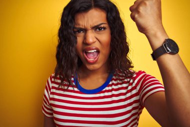 Eterseksüel transeksüel kadın izole sarı arka plan üzerinde stiped t-shirt giyen kızgın ve öfke, deli ve yükseltilmiş el, öfke kavramı ile bağırarak sinirli