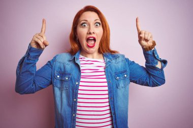 Denim gömlek giyen güzel kızıl saçlı kadın ve izole pembe arka plan üzerinde çizgili t-shirt gülümseyerek şaşırttı ve şaşırttı ve parmakları ile işaret ve kolları kaldırdı.
